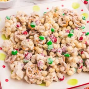 White trash candy (white chocolate chex mix candy) on a plate surrounded by small bowls of peanuts, cereal, pretzels, and Christmas colored M&Ms.