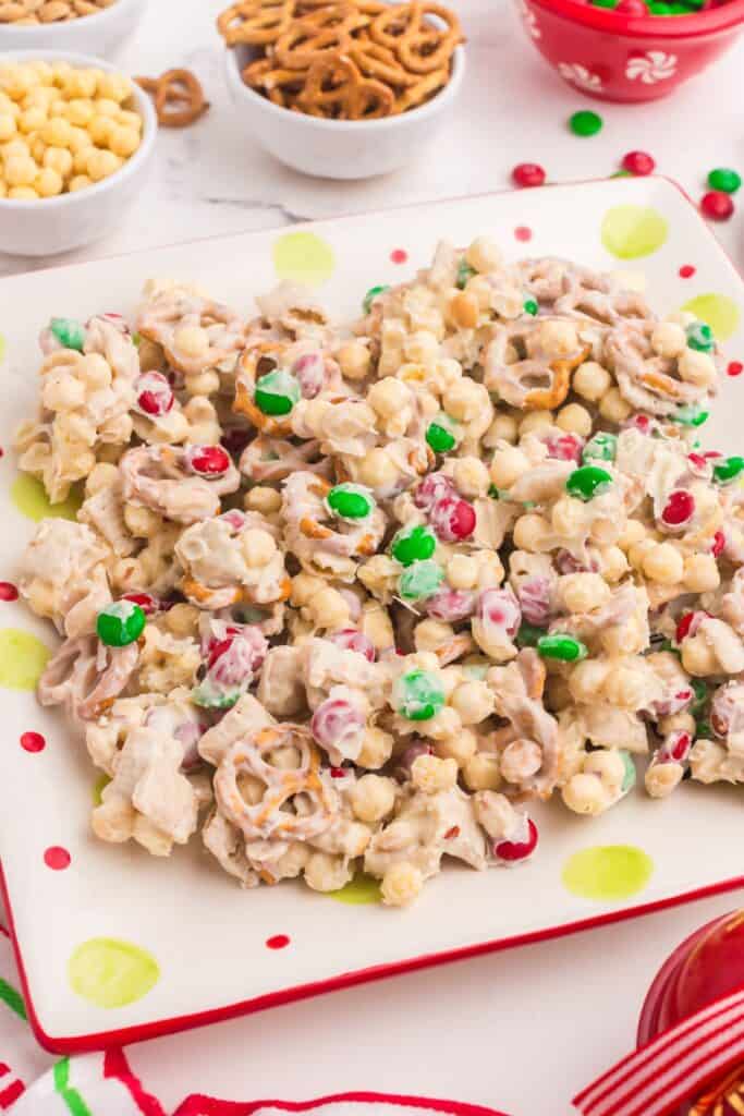 White trash candy (white chocolate chex mix candy) on a plate surrounded by small bowls of peanuts, cereal, pretzels, and Christmas colored M&Ms.