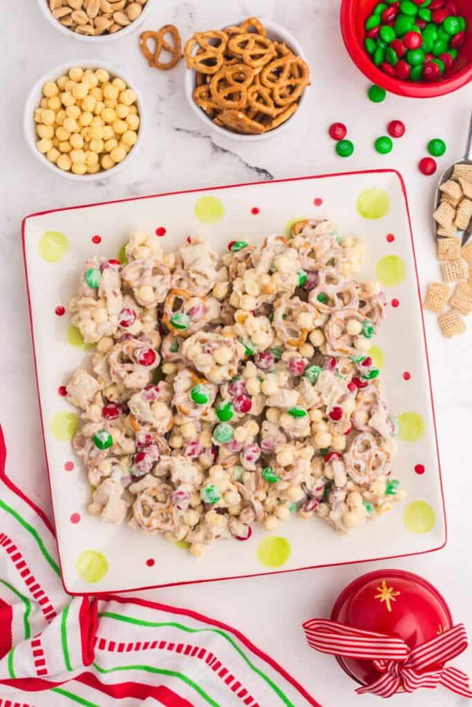 White trash candy (white chocolate chex mix candy) on a plate surrounded by small bowls of peanuts, cereal, pretzels, and Christmas colored M&Ms.