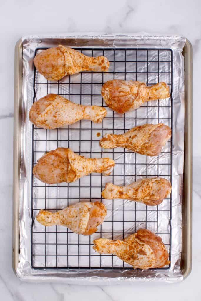 Overhead view of seasoned chicken legs before baking on a cooling rack, over an aluminum foil lined baking sheet.