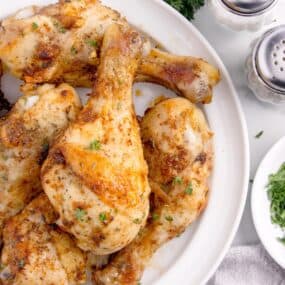 White serving plate with baked chicken legs on it. Surrounded by fresh parsley, and salt and pepper shakers.