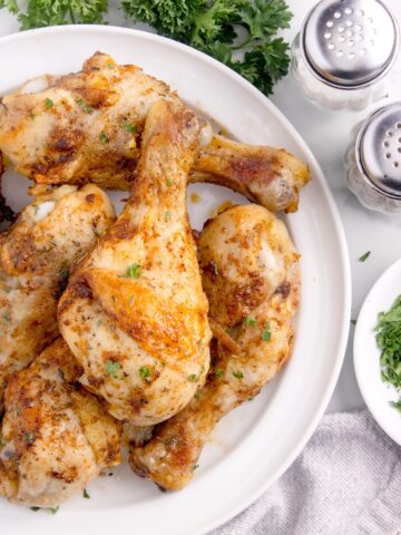 White serving plate with baked chicken legs on it. Surrounded by fresh parsley, and salt and pepper shakers.