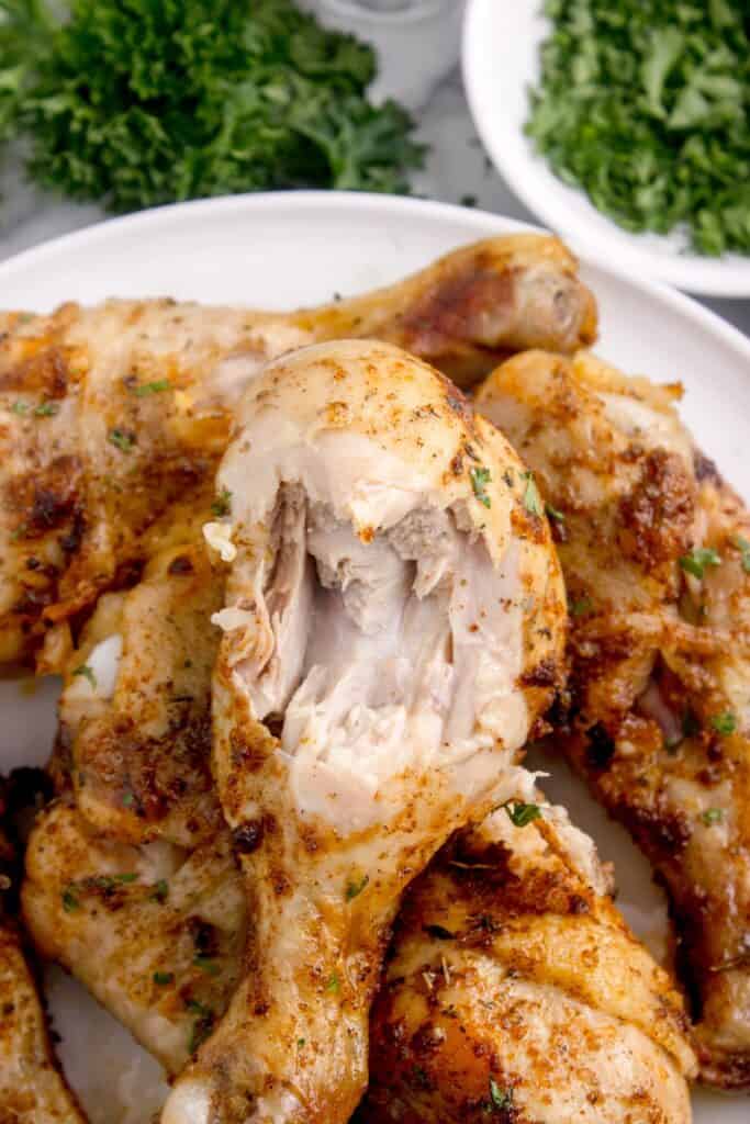 Chicken legs piled up on a white dinner plate. The top piece of chicken has a bite taken out of it to reveal the juicy center.