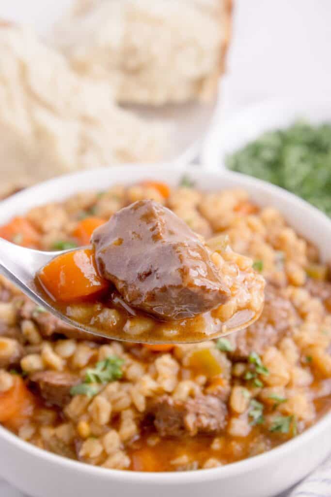 A spoon has elevated a bite of beef and barley soup about a full soup bowl.