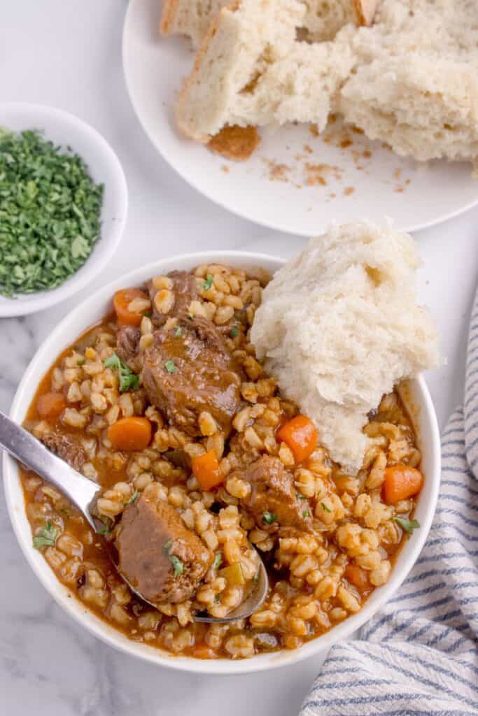 Overhead view of a white soup bowl with beef and barley soup with a hunk of bread and parsley garnish.