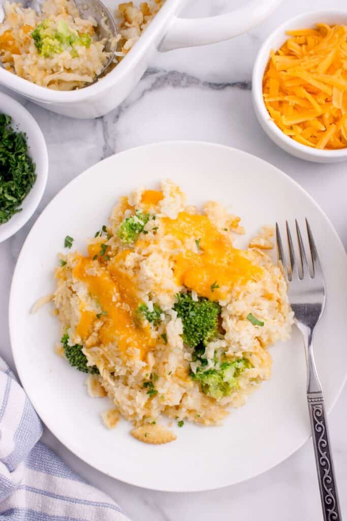 A white dinner plate with a serving of chicken broccoli rice casserole and a fork. Small bowl of shredded cheese and small bowl of parsley are above the plate.