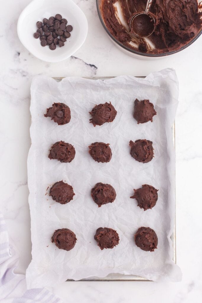 Unbaked cookie dough on a parchment lined cookie sheet. Bowl of cookie dough with a cookie scoop above the cookie sheet.