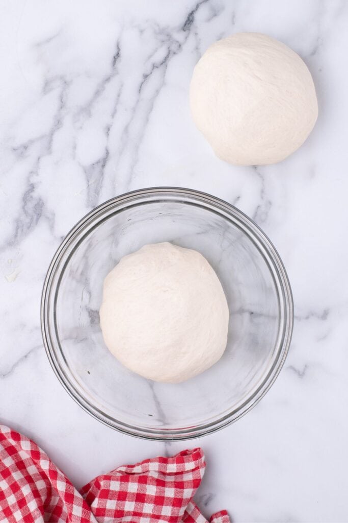 Pizza dough divided into two round balls. One is in a glass bowl, the other on the table top.