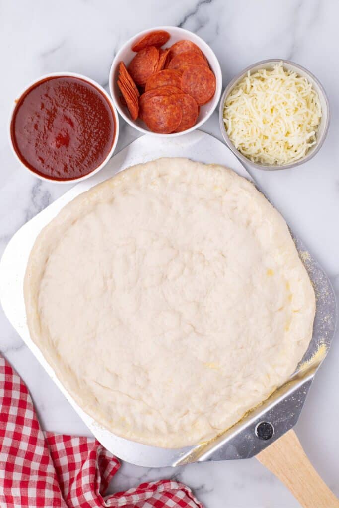 Easy pizza crust recipe on a pizza peel. Three bowls of toppings (red sauce, pepperoni, and mozzarella cheese) sit just above the peel.