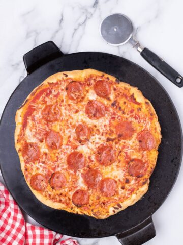 Large baking stone with a baked pizza on it. Pizza has red sauce, cheese, and pepperoni. Pizza cutter sitting just above the pizza.