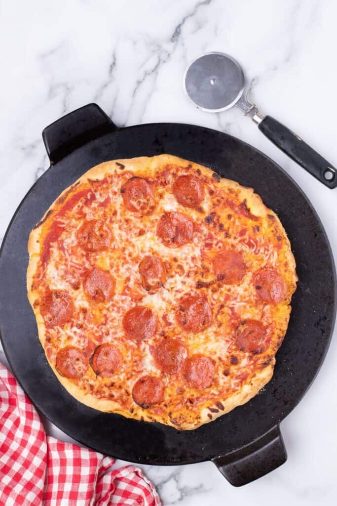 Large baking stone with a baked pizza on it. Pizza has red sauce, cheese, and pepperoni. Pizza cutter sitting just above the pizza.