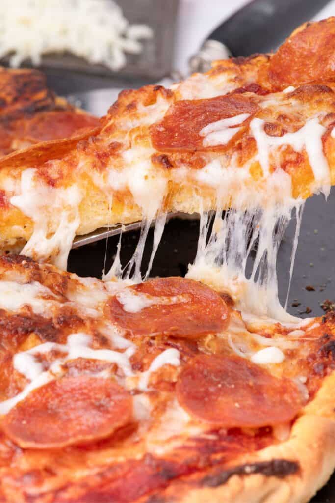 Side view of a slice of pizza being lifted away from the rest of the pie. Shows cross section of the crust and a cheese pull.