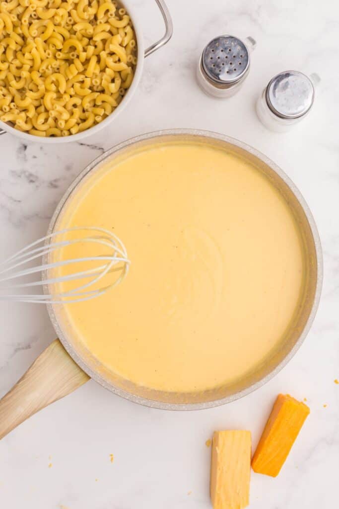 Cheese sauce whisked in a large skillet. Bowl of cooked elbow noodles above in a pot.