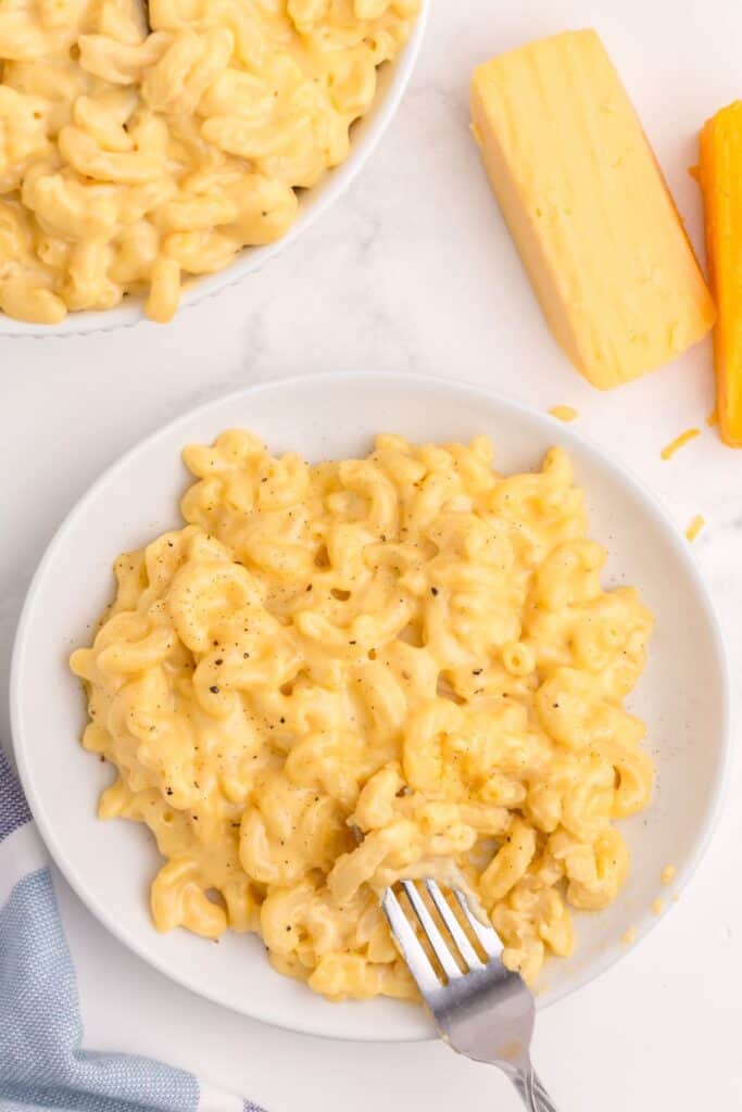 White dinner plate and fork with gouda mac and cheese on it. 2 small blocks of cheese are above the plate.