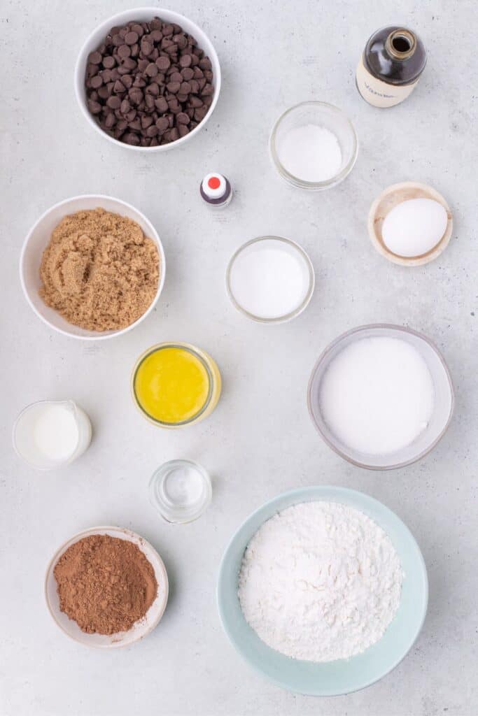 Ingredients for red velvet cookies: flour, cocoa powder, baking soda, salt, brown and white sugar, butter, egg, whole milk, vanilla extract, white vinegar, red gel food coloring, and chocolate chips.