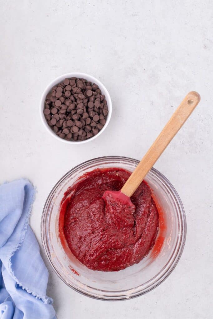 Glass bowl of red velvet cookie dough batter with a bowl of chocolate chips above it.