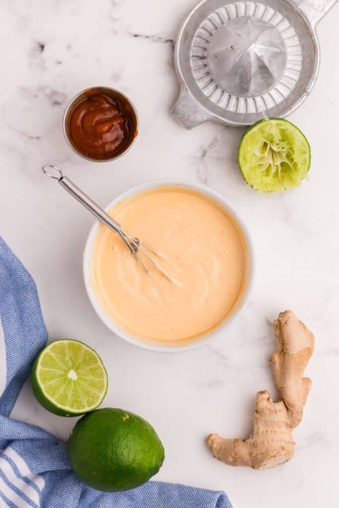 Bowl of mayo with a small whisk surrounded by limes, ginger, a citrus juicer, and Sriracha.