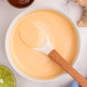 Close up of a bowl of mayo. Small condiment bowl of Sriracha, ginger, and limes surrounding the bowl.