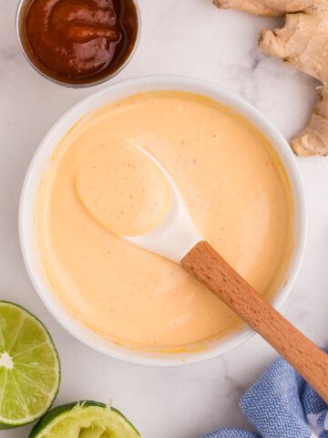 Close up of a bowl of mayo. Small condiment bowl of Sriracha, ginger, and limes surrounding the bowl.