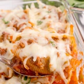 Casserole dish with beef shells. A serving spoon is lifting up a portion of the recipe.