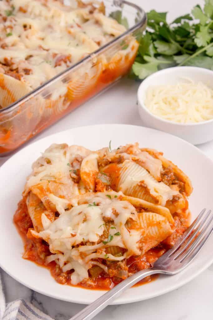 White dinner plate with beef shells and a fork. Casserole dish of shells and a bowl with shredded parmesan is in the background.