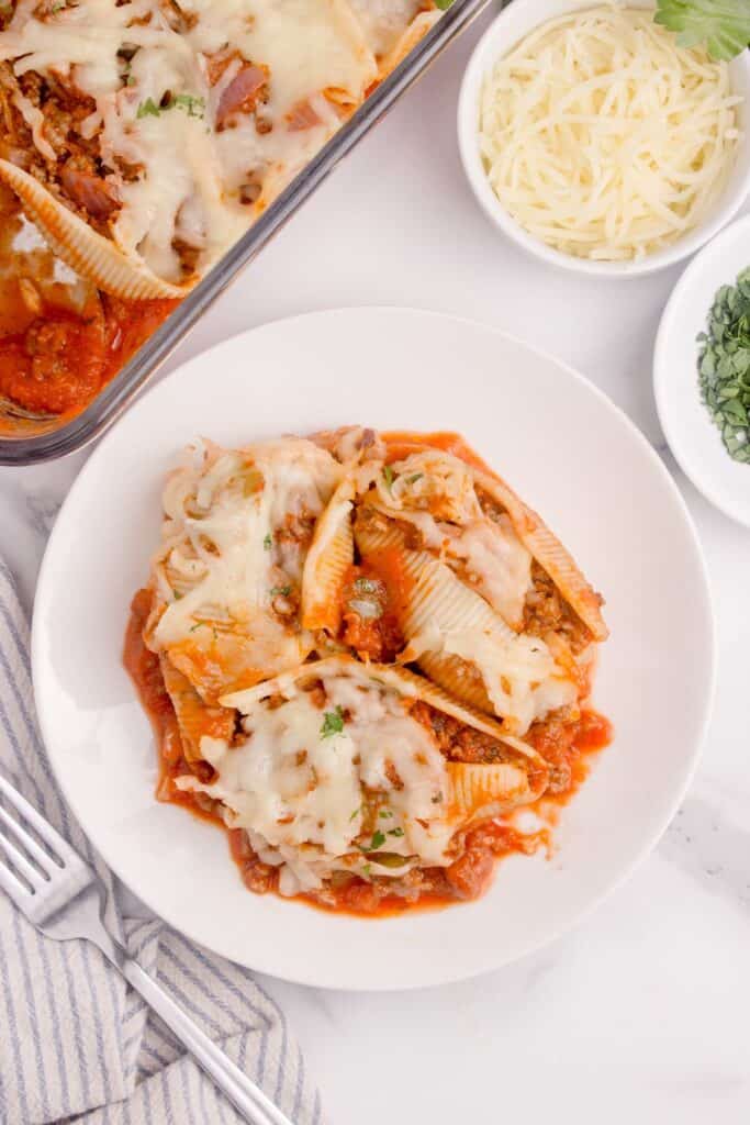 Overhead view of a white dinner plate with stuffed shells on it. Plate is surrounded by the casserole dish of shells, a fork, and two bowls of shredded parmesan cheese, and chopped parsley.