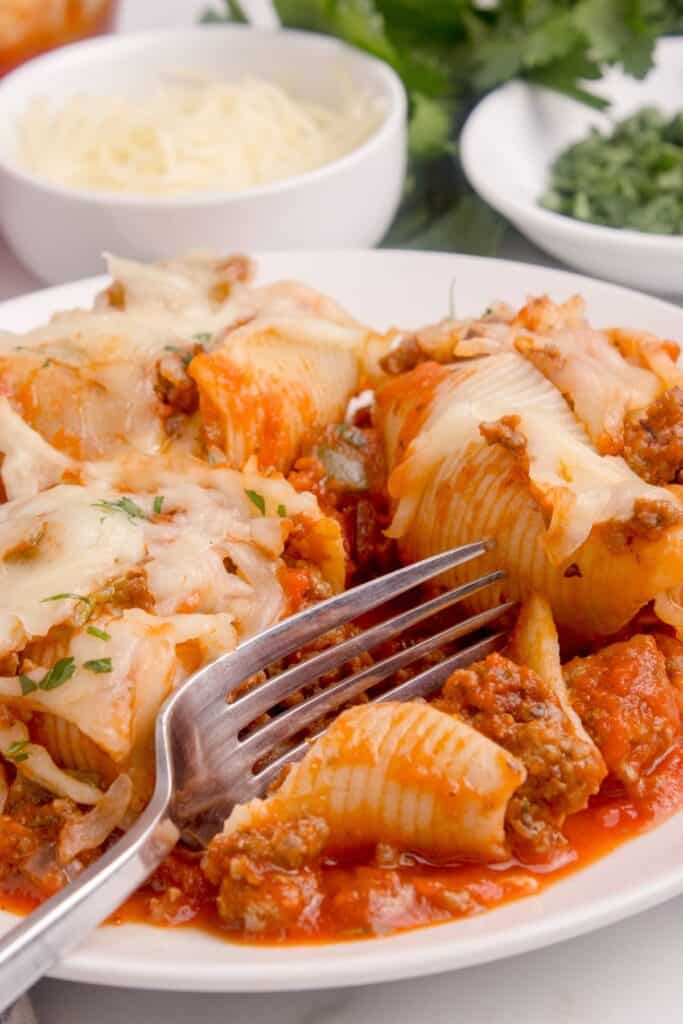 White dinner plate with beef stuffed shells and a fork. Two small bowls in the background filled with shredded parmesan cheese and chopped parsley.