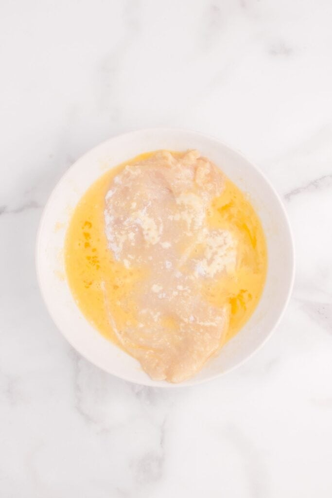 Raw chicken in a bowl of beaten eggs. Chicken is being prepped for breadcrumb coating.