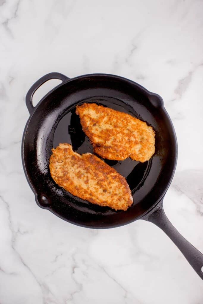 Cast iron pan with two chicken cutlets cooking.