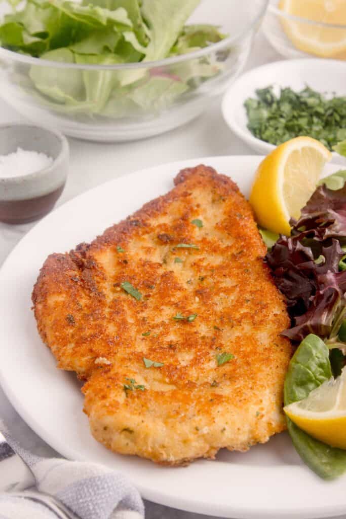 Chicken cutlet on a white dinner plate with a green salad and lemon wedge.