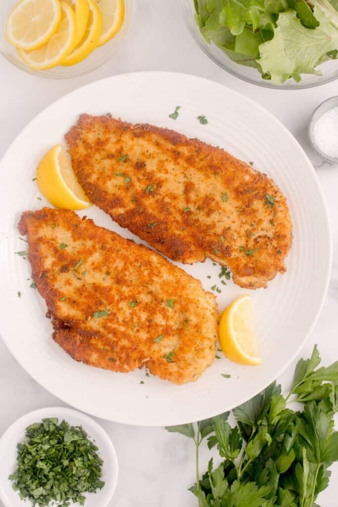 White dinner plate with two breaded chicken cutlets and lemon wedges on it.