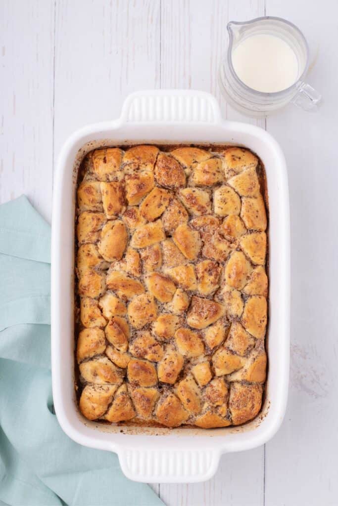 Baked casserole in a white baking dish. Pitcher of cinnamon roll glaze just next to the casserole.