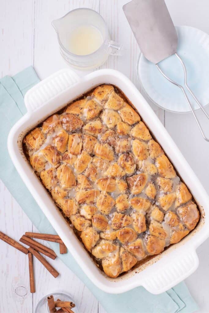 Completed casserole surrounded by serving plates, a pitcher of glaze, and cinnamon sticks.