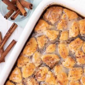 Casserole fresh out of the oven with a glaze drizzled over it. Cup of cinnamon sticks sits just above the baking dish.