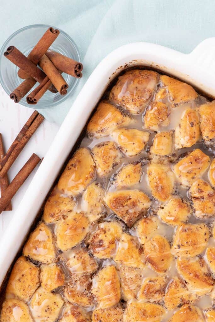 Casserole fresh out of the oven with a glaze drizzled over it. Cup of cinnamon sticks sits just above the baking dish.