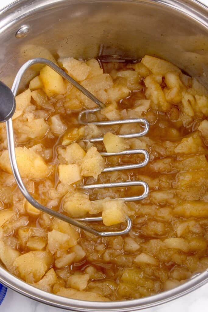 Pot of cooked apples being mashed with a potato masher.