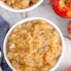 Two bowls of chunky cinnamon applesauce, a silver spoon, and whole apples.