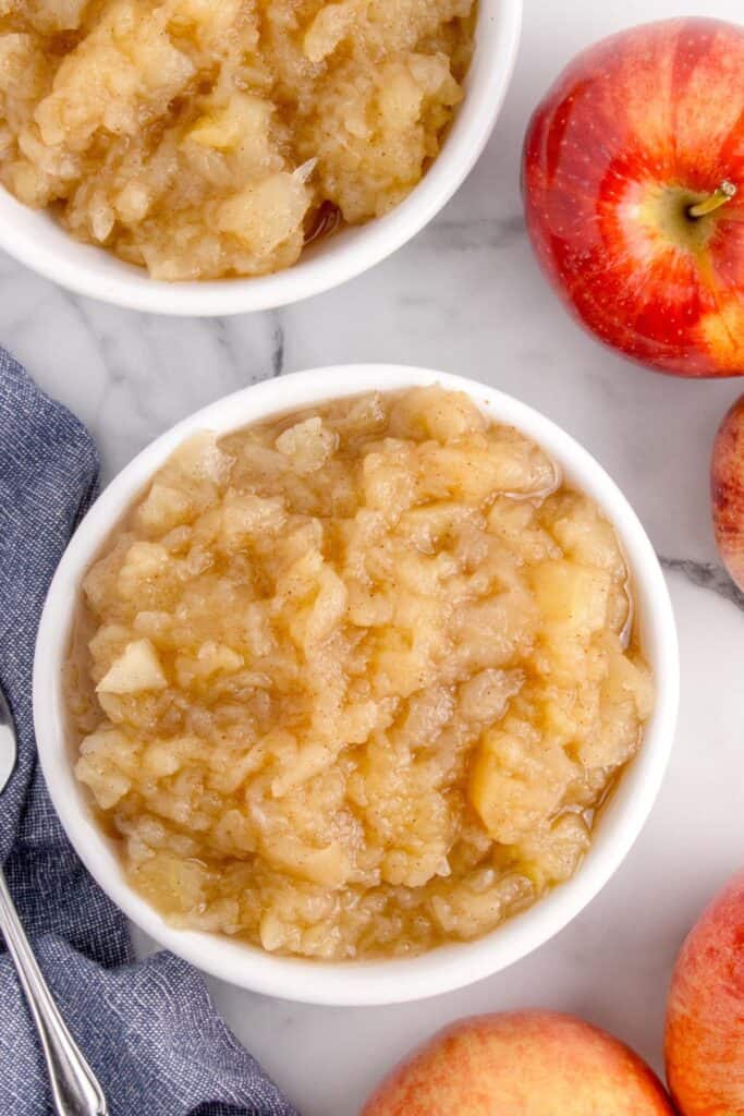 Two bowls of chunky cinnamon applesauce, a silver spoon, and whole apples.