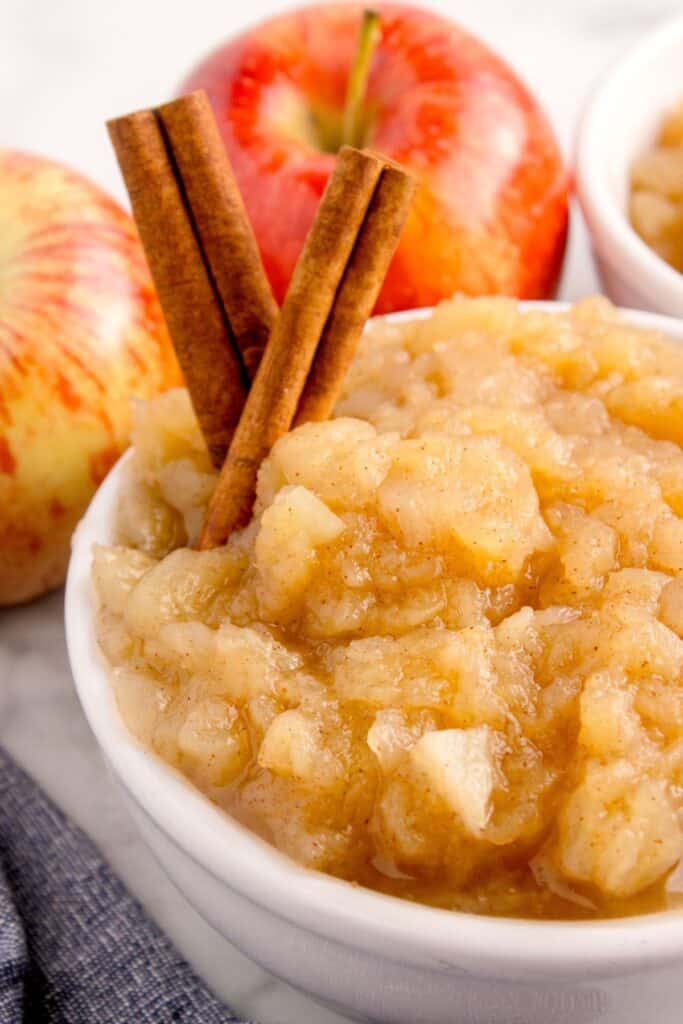 A white bowl of cinnamon applesauce with a cinnamon stick garnish. Whole apples in the background.