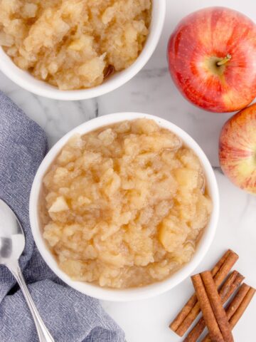 Two bowls of chunky cinnamon applesauce, a silver spoon, cinnamon sticks, and whole apples.