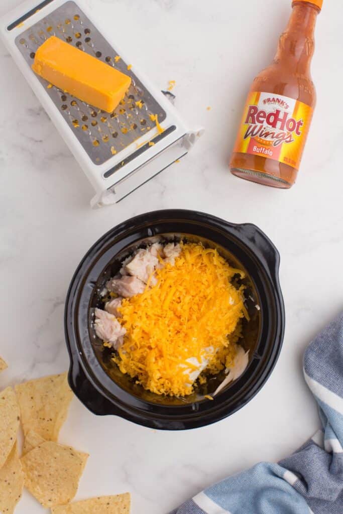 Buffalo chicken dip assembly. Chicken at the bottom of the crock pot with shredded cheese on top.