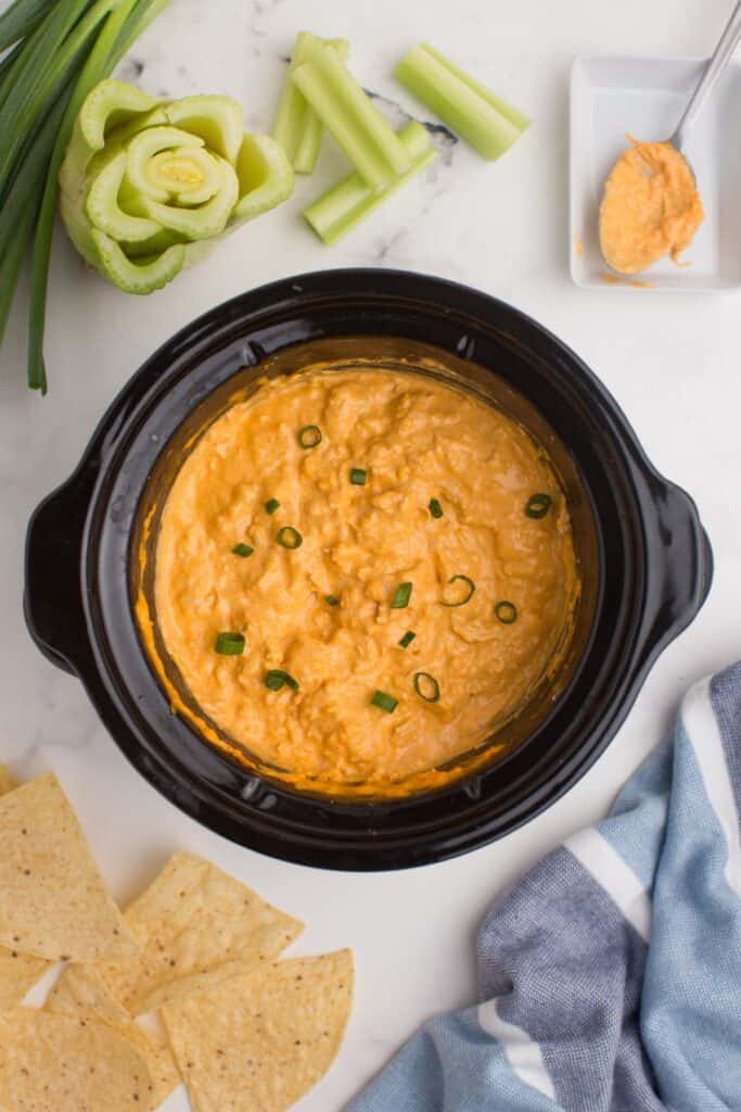 Buffalo chicken dip in a small slow cooker surrounded by tortilla chips, green onions, celery stalks, and a plate with a spoon.