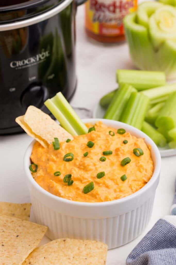 Serving bowl of dip surrounded by tortilla chips, celery, a bottle of hot sauce, and a crockpot.