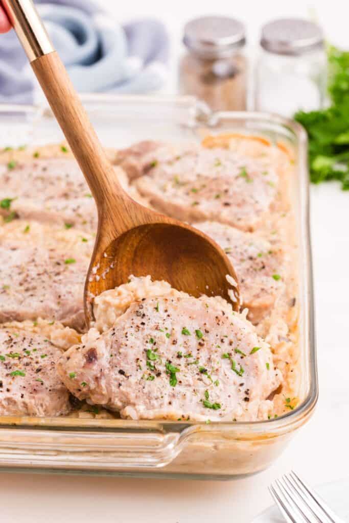 Glass baking dish with pork chop casserole. Wooden spoon is lifting out a serving.