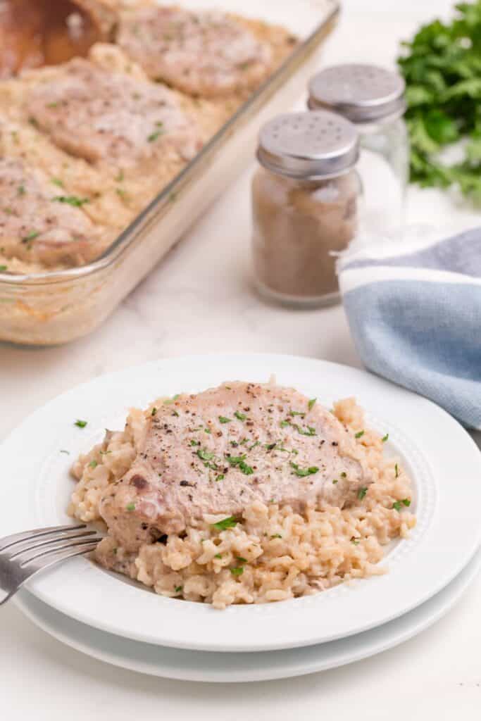 Stacked white dinner plates with a serving of pork chop casserole on the top dish. Glass casserole dish in the background with fresh parsley and salt and pepper shakers.