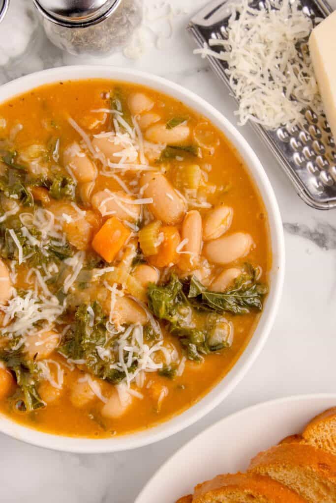 Bowl of white bean soup surrounded by a plate of toast, a block of parmesan cheese, a cheese grater, a soup spoon, and salt and pepper shakers.