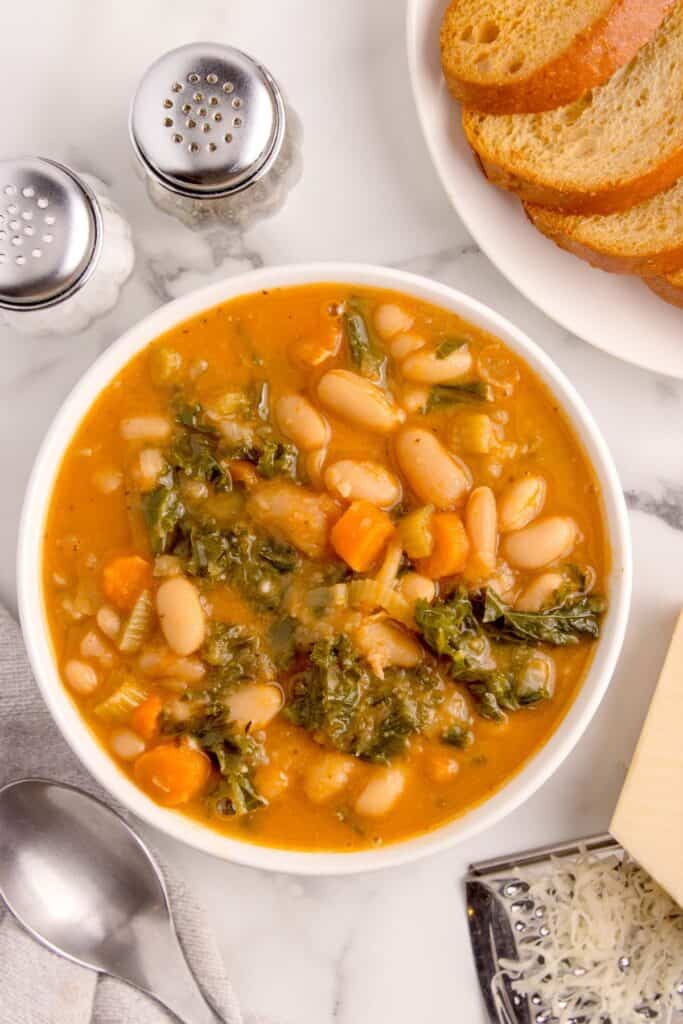 Bowl of white bean soup surrounded by a plate of toast, a block of parmesan cheese, a cheese grater, a soup spoon, and salt and pepper shakers.