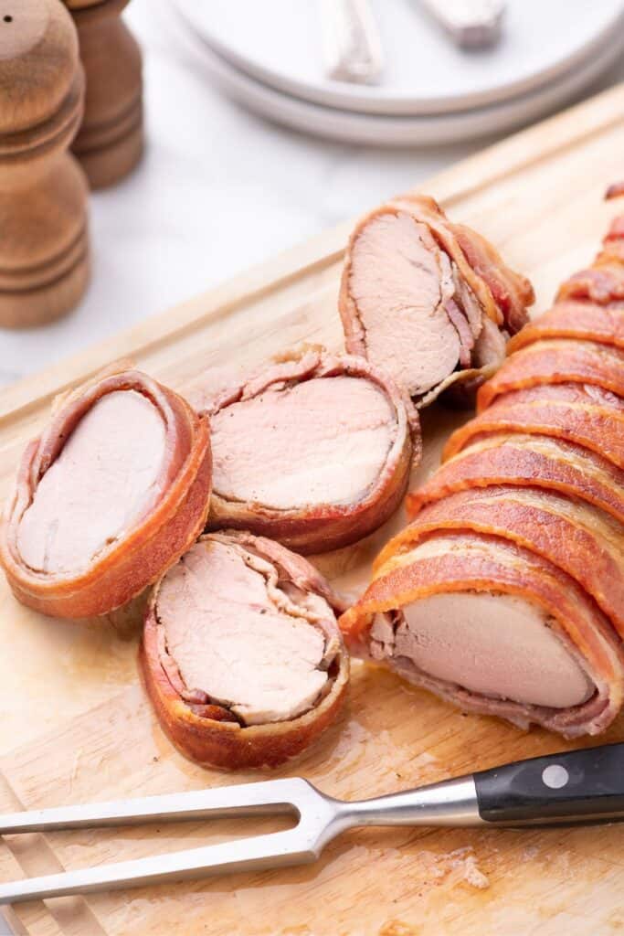 Large cutting board with a carving fork and a sliced bacon wrapped pork tenderloin on it.