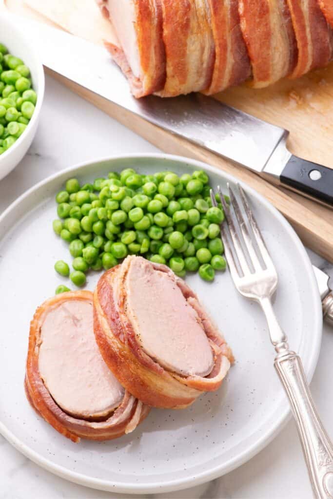 A white dinner plate with two slices of bacon wrapped pork tenderloin and a serving of green peas with a fork on the plate.