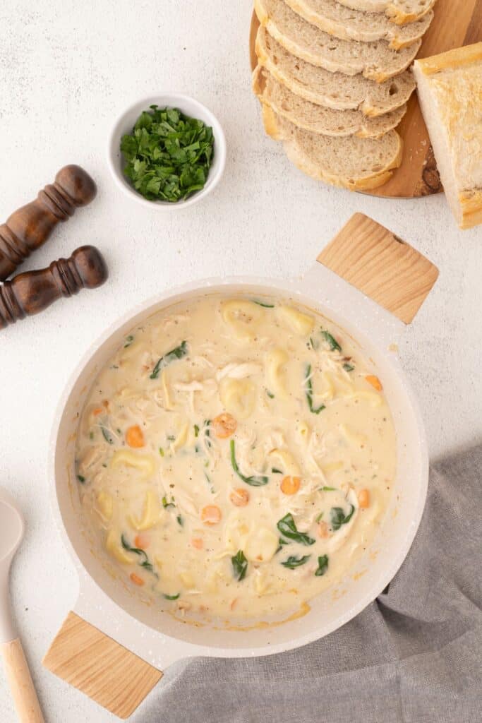 Pot of chicken tortellini soup surrounded by sliced bread on a cutting board, parley, and salt and pepper shakers.